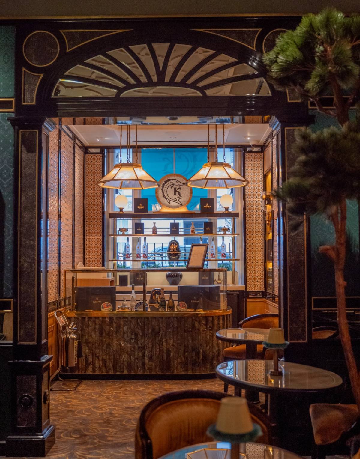 A stylish, dimly-lit bar with an ornate, arching entrance. The bar counter features an array of liquor bottles and a clock in the centre. Elegant pendant lights hang above, illuminating the cosy seating area with plush chairs and small tables.