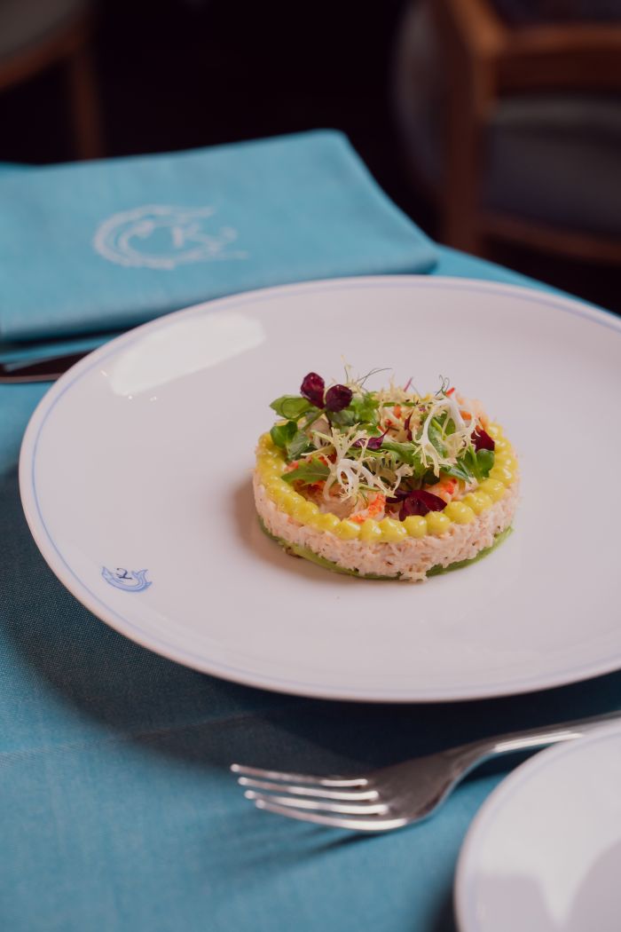 A gourmet dish is elegantly presented on a white plate with a blue rim, featuring a circular arrangement of artfully plated vegetables and garnishes. The plate rests on a light blue tablecloth, with a napkin bearing an emblem placed nearby. A fork is visible in the foreground.