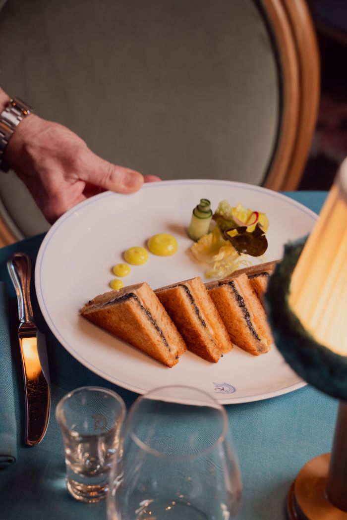 A hand places a white plate with three sandwich triangles, yellow sauce, and a vegetable garnish on a table set for dining with cutlery, a lamp, and glassware.