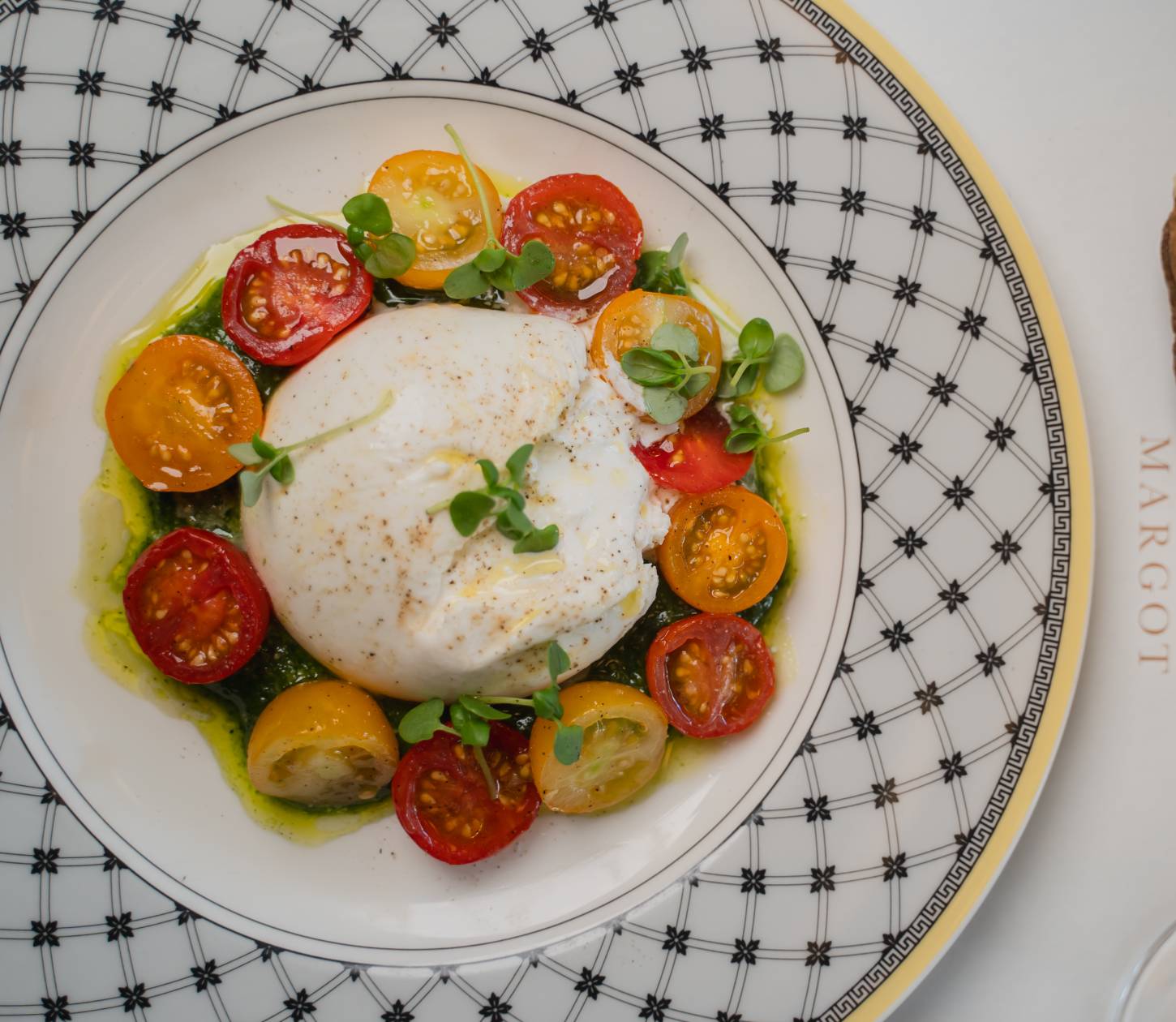 A dish featuring a creamy burrata cheese surrounded by colorful tomato slices and garnished with microgreens. The plate has a patterned black and white design. Next to it, there's a piece of focaccia bread with a menu partially visible underneath.