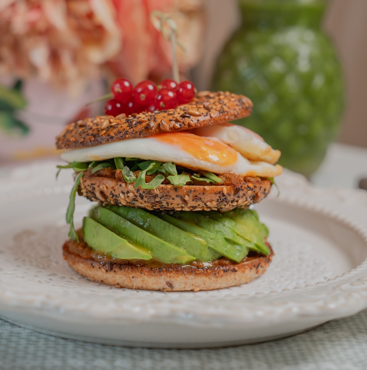 A gourmet sandwich on a white plate features a seeded bagel, layered with sliced avocado, arugula, a fried egg with a runny yolk, and red currants on top. The setting includes blurred background elements like a green vase and pink flowers, adding a rustic touch.