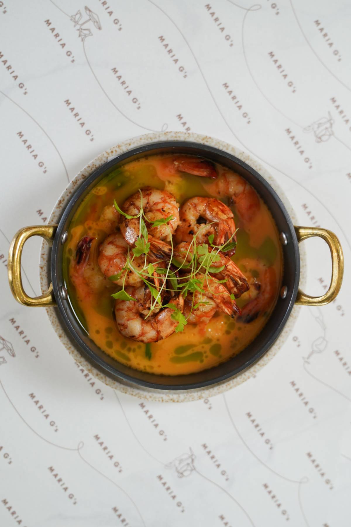 A bowl of shrimp in a yellow broth garnished with microgreens. The dish is served in a round, handled pot with golden handles on a white tablecloth adorned with repeating patterns.